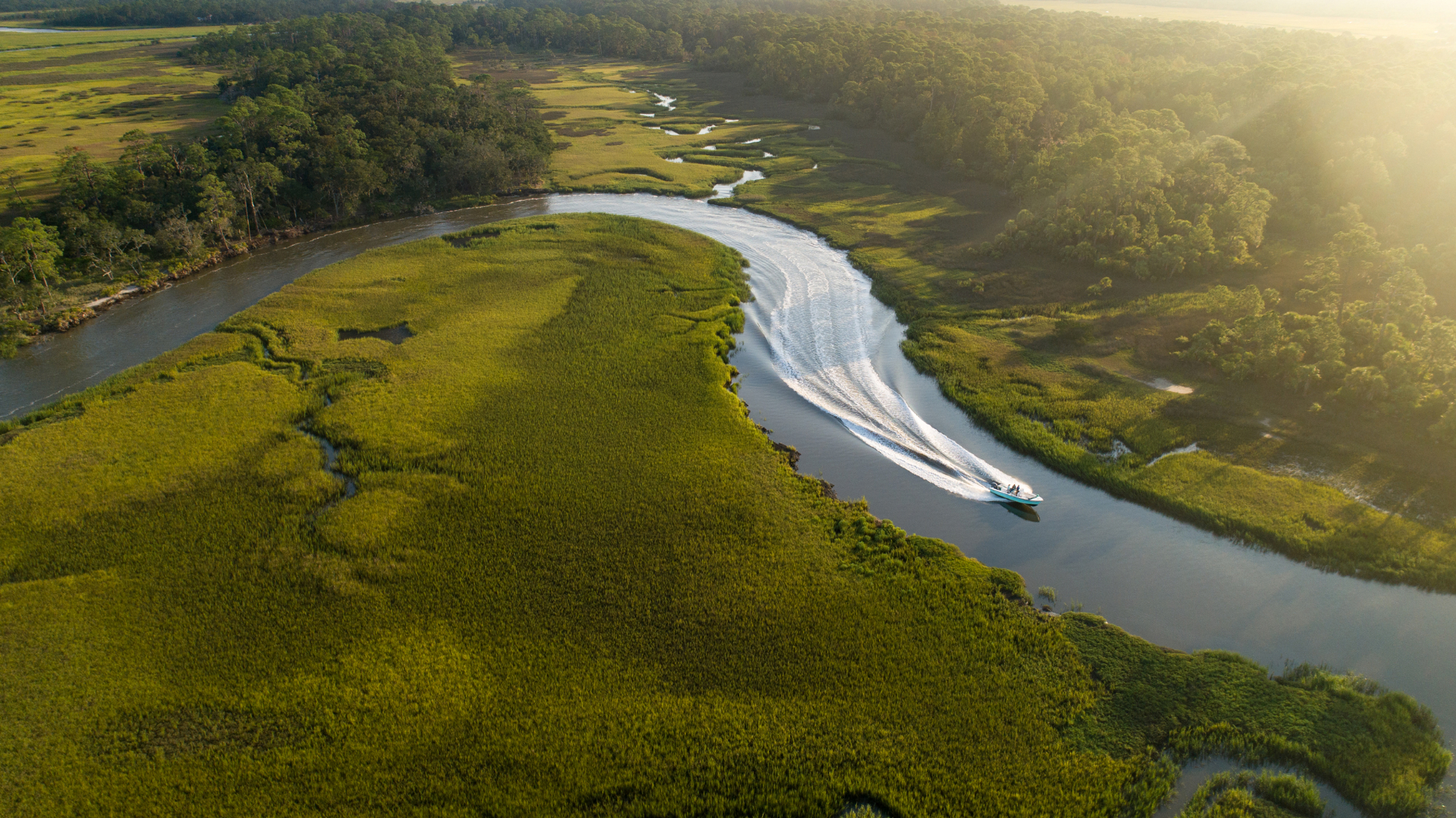 Aerial view of a winding river through sunlit marshland with a boat leaving a silver wake